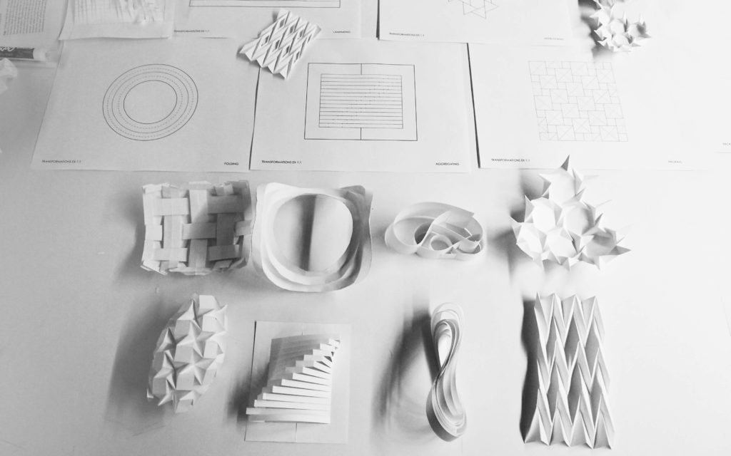 Image of folded paper three-dimensonal models on a studio desk with drawings showing the precise dimensioning of paper that constructed them.  