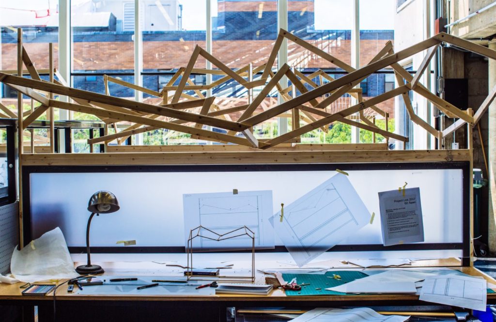Image of a studio desk inside Gund Hall at the Harvard Graduate School of Design.  On the desk is a desk lamp, a partially build model out of balsa wood framing, several drawings, and large timber canopy over the desk.  