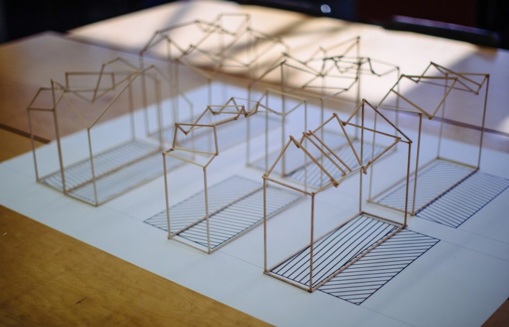 Image of six study models in thin wood sticks defining the corners and edges of multi-story houses with different gable roof profiles.  The models are sitting on a studio desk on top of a paper printed with a striped pattern.  